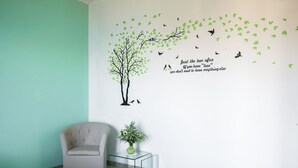 Une pièce avec un mur de couleur turquoise, un mur blanc orné d’un motif d’arbre et d’une citation, un fauteuil beige et une table en verre transparent sur laquelle est posé un vase.