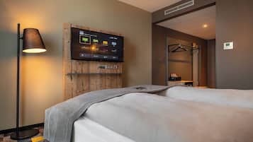 A hotel room with a bed, a TV mounted on a wooden panel, a floor lamp, and a mirror.