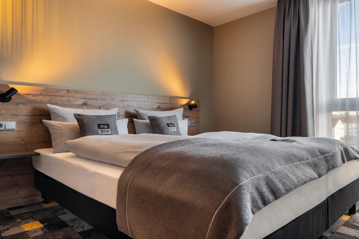 A modern hotel room with a wooden headboard, a large bed with multiple pillows, a bedside table, and a view of the city through large windows.