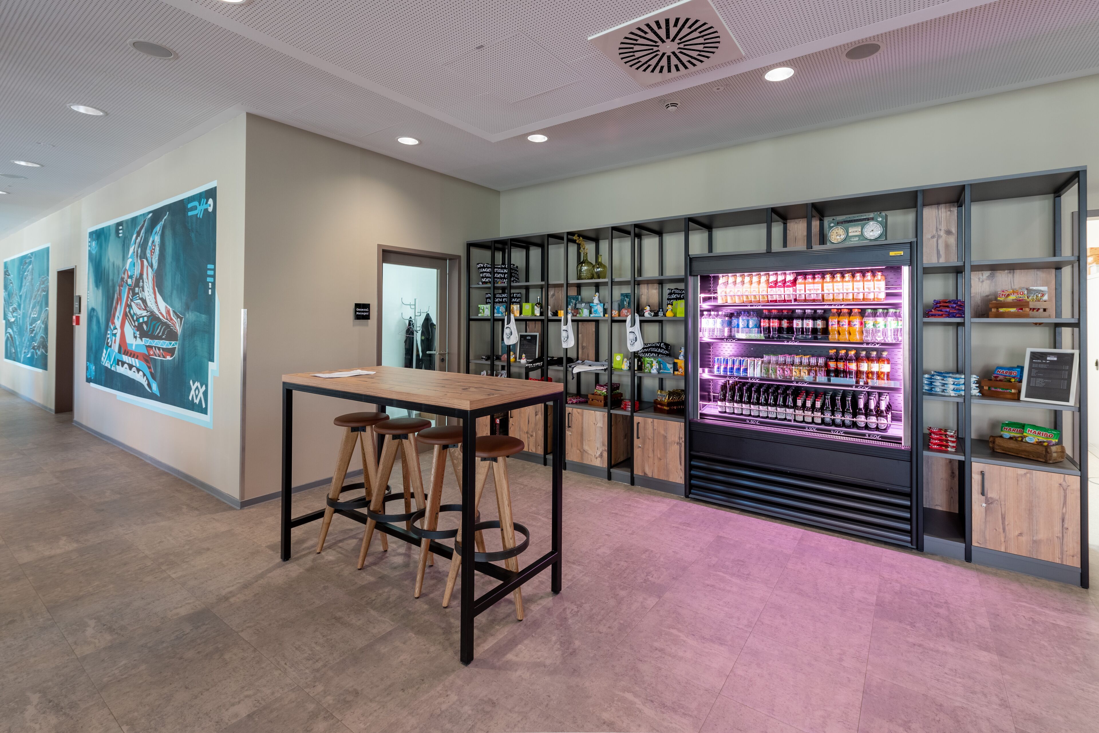 A modern bar area with a wooden table and stools, a neon-lit beverage dispenser, and shelves with various bottles and snacks.