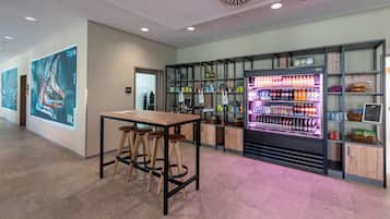 Un moderno bar con una mesa de madera y taburetes, un dispensador de bebidas iluminado con neón, y estantes con varias botellas y aperitivos.