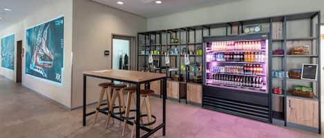 A modern bar area with a wooden table and stools, a neon-lit beverage dispenser, and shelves with various bottles and snacks.