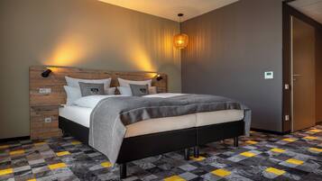 A modern hotel room with a wooden headboard, a bed with grey bedding, a patterned floor, and a wall-mounted light fixture.