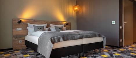 A modern hotel room with a wooden headboard, a bed with grey bedding, a patterned floor, and a wall-mounted light fixture.