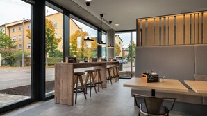 A modern cafe with wooden furniture, large windows, and a view of the street. - Holiday Inn - the niu, Timber Esslingen by IHG (Esslingen)