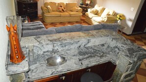Una cocina con encimera de mármol, pileta y taburetes de bar. Se ve un sofá y una televisión al fondo.