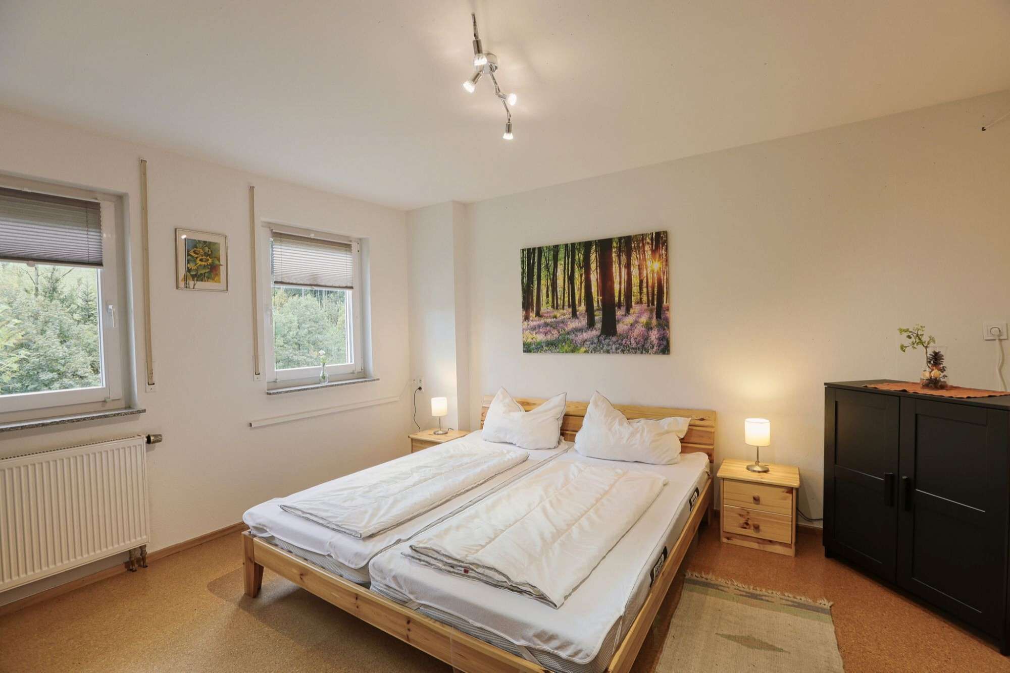 A bedroom with two beds, a radiator, a window with blinds, a painting, a bedside table with a lamp, and a wardrobe.