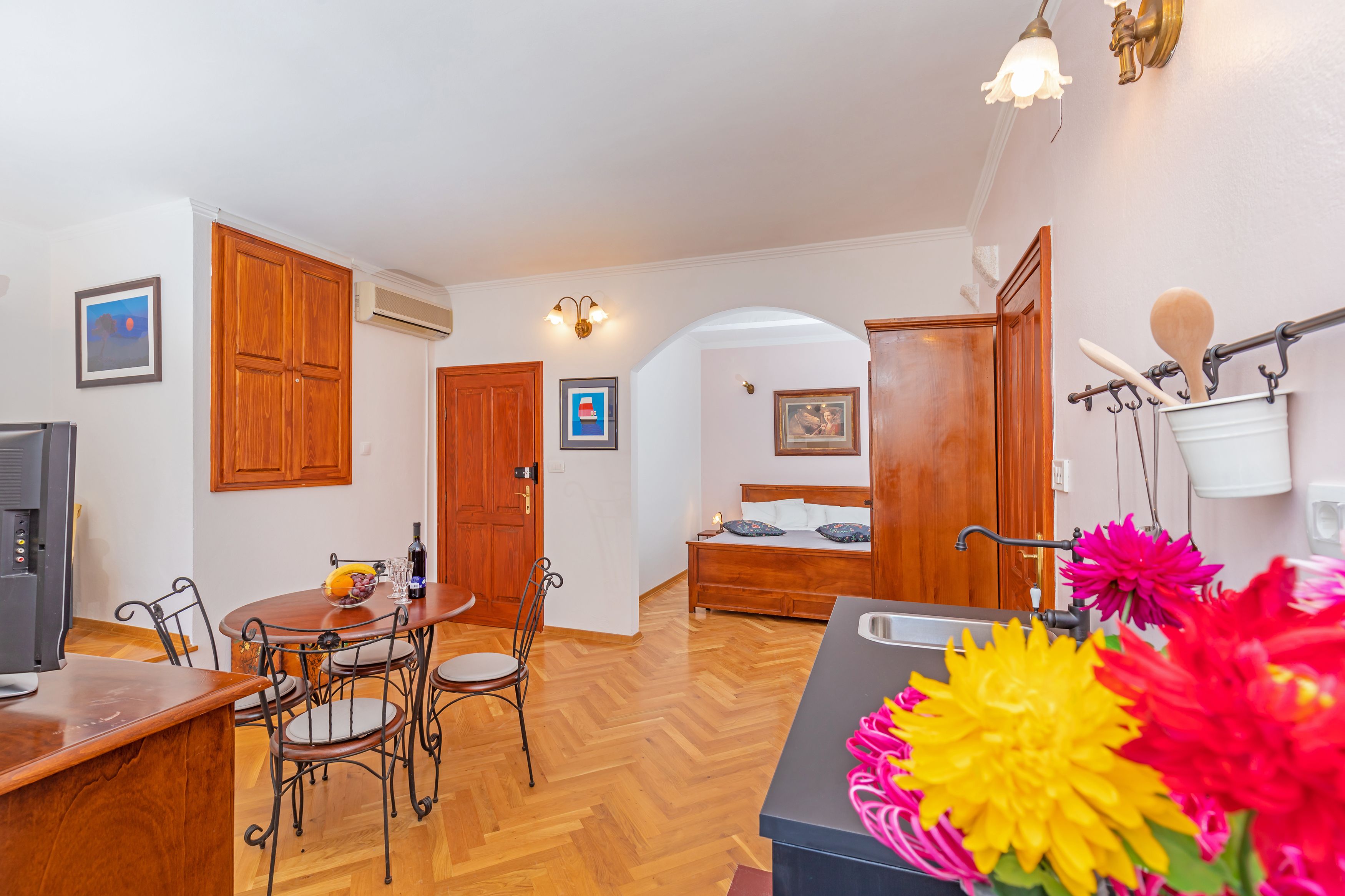 A compact hotel room with a wooden dining table, chairs, a bed, and a kitchenette.