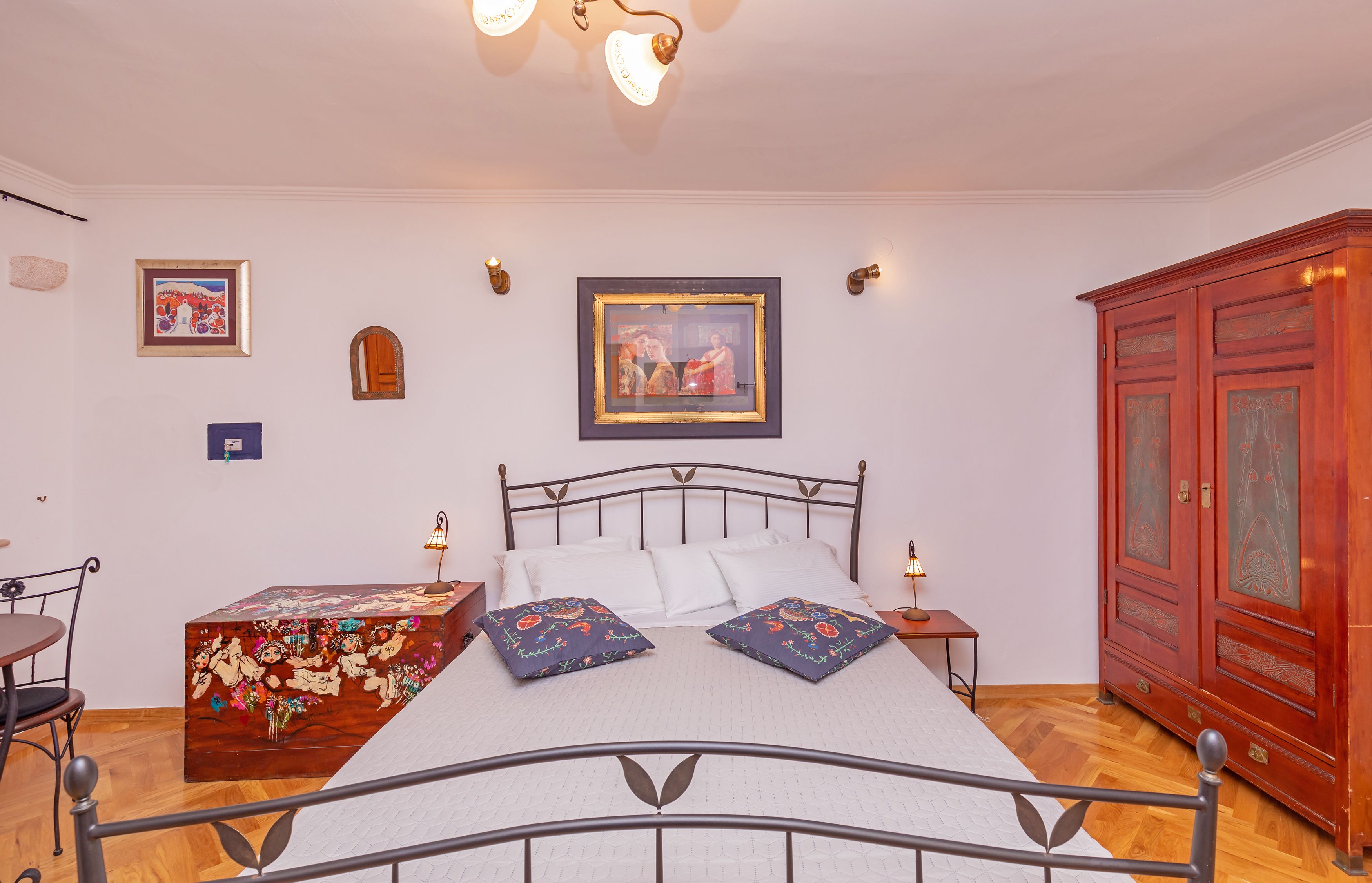 A bedroom with a metal bed frame, white bedding, two decorative pillows, a wooden chest of drawers, a red wardrobe, and two wall-mounted lamps.