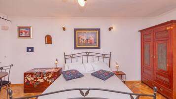 A bedroom with a metal bed frame, white bedding, two decorative pillows, a wooden chest of drawers, a red wardrobe, and two wall-mounted lamps.