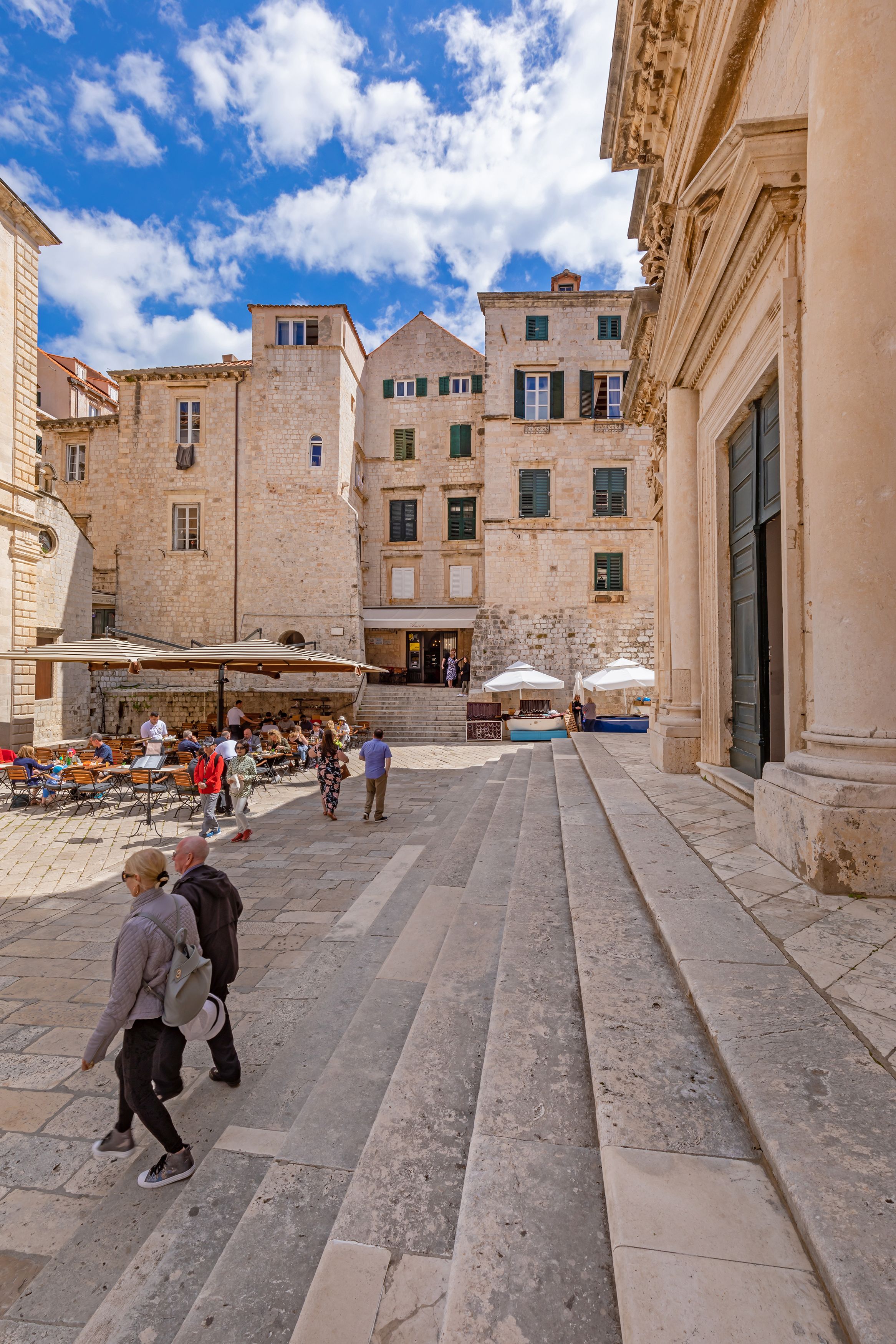 A courtyard with stone buildings, outdoor seating, and a staircase leading to an upper floor.