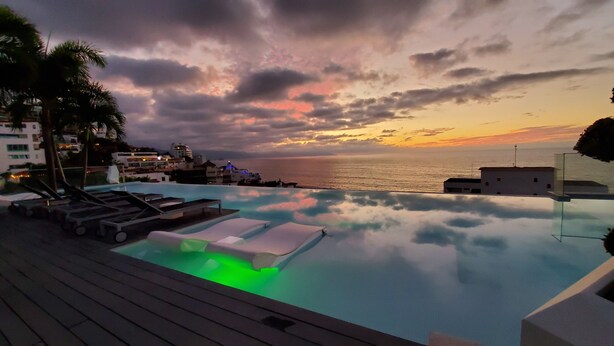 En infinitypool med solstolar, ett trädäck och utsikt över havet vid solnedgången.