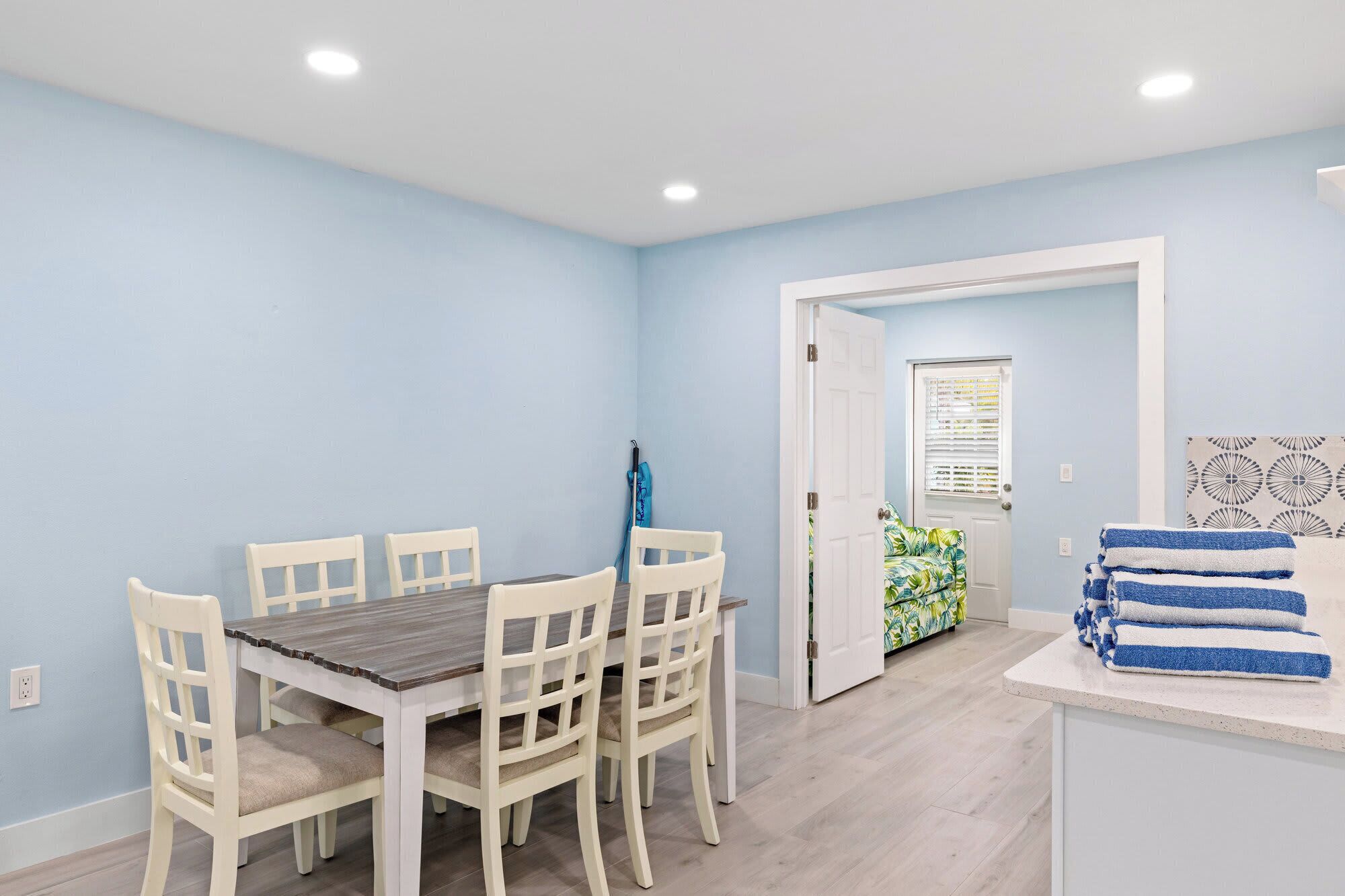 Un coin repas avec une table en bois et des chaises blanches, une porte donnant sur une chambre, et une pile de serviettes sur un comptoir.