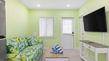 Una habitación con pared verde, un sofá estampado con motivos tropicales, una mesa de madera con toallas, un televisor en la pared y una puerta blanca.