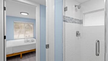 A bathroom with a glass shower enclosure, a built-in bench with a white mattress, and a window with blinds.