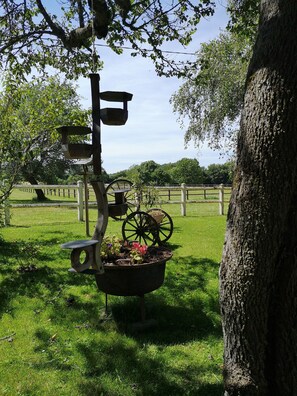 Un espacio al aire libre rústico con una escultura de metal que presenta una rueda y elementos colgantes, rodeado de exuberante vegetación y una valla de madera.