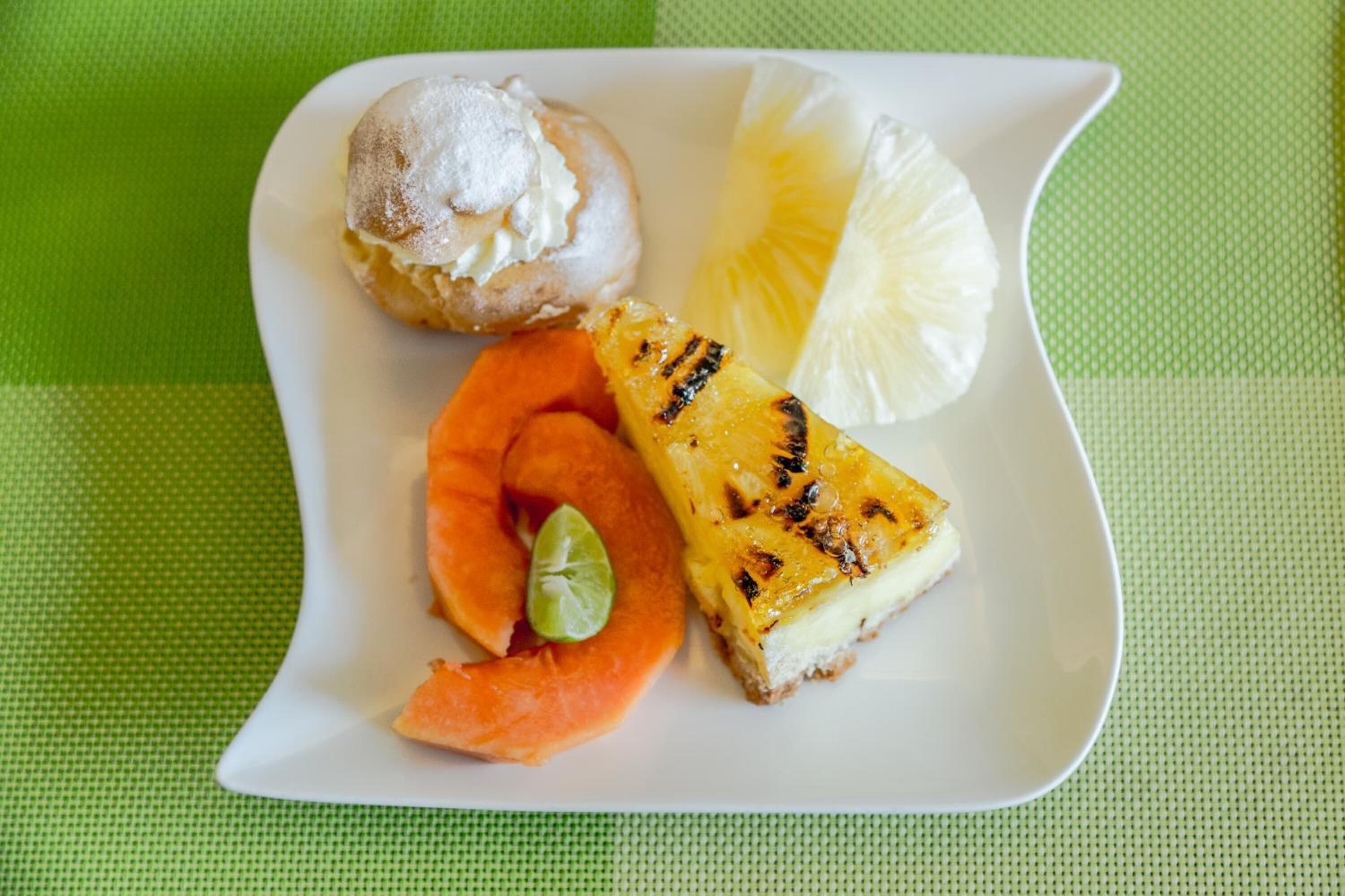 A plate with a pastry, a slice of pineapple, a slice of cheesecake, and a lime wedge.