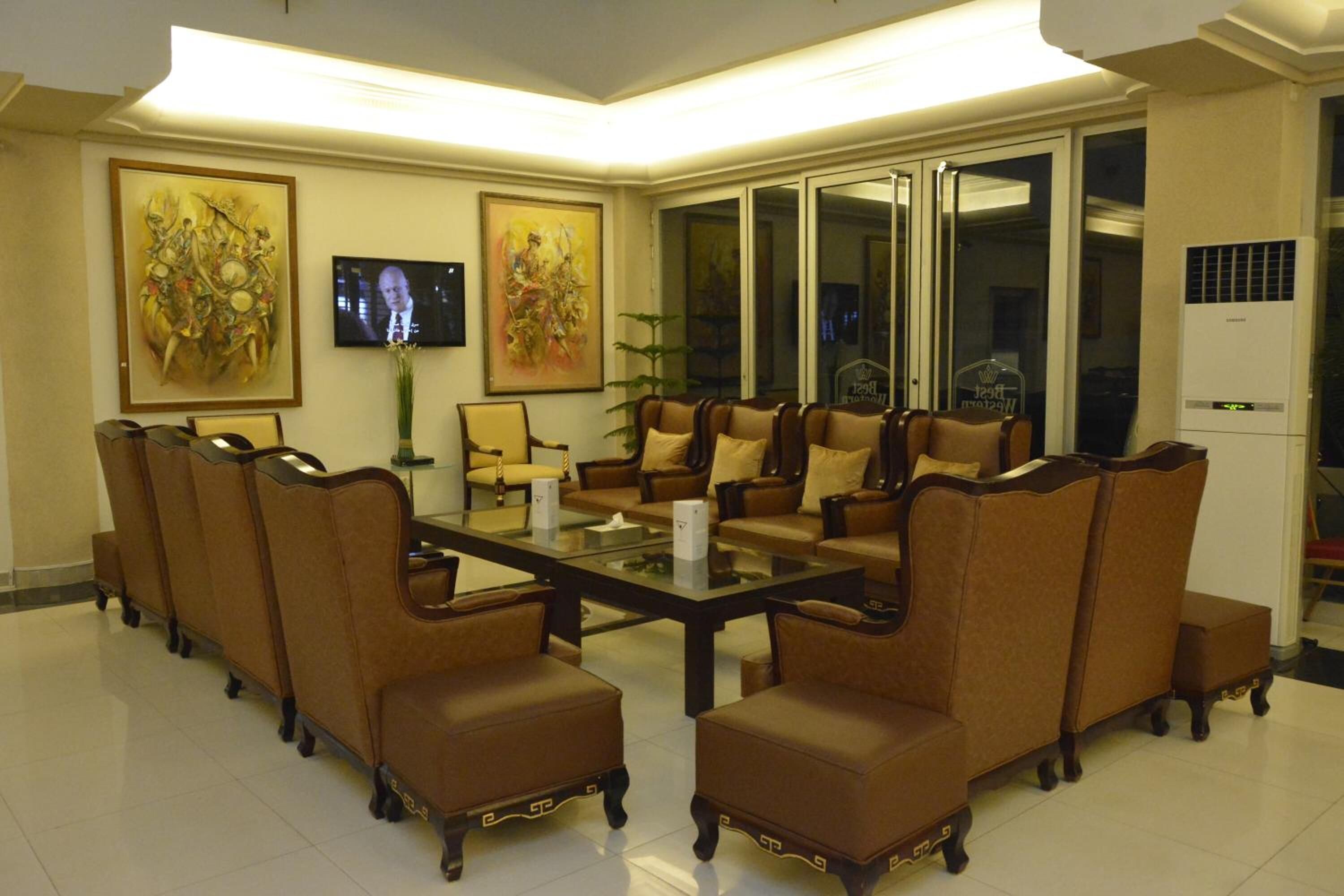 A conference room with a glass table surrounded by brown upholstered chairs, two large paintings on the walls, and a television screen.