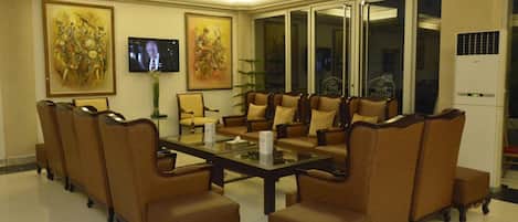 A conference room with a glass table surrounded by brown upholstered chairs, two large paintings on the walls, and a television screen.