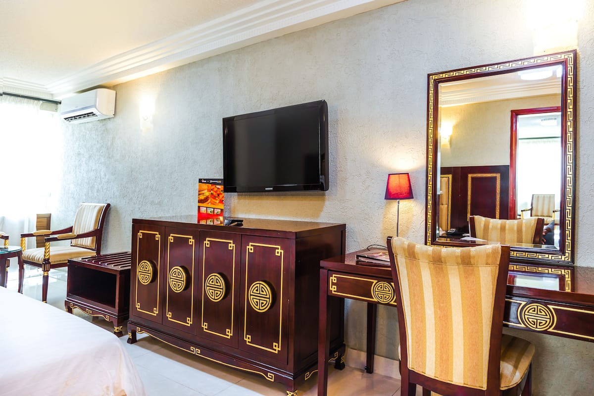 A hotel room with a flat-screen TV mounted on the wall, a wooden cabinet with decorative panels, a striped armchair, and a large mirror with a decorative frame.