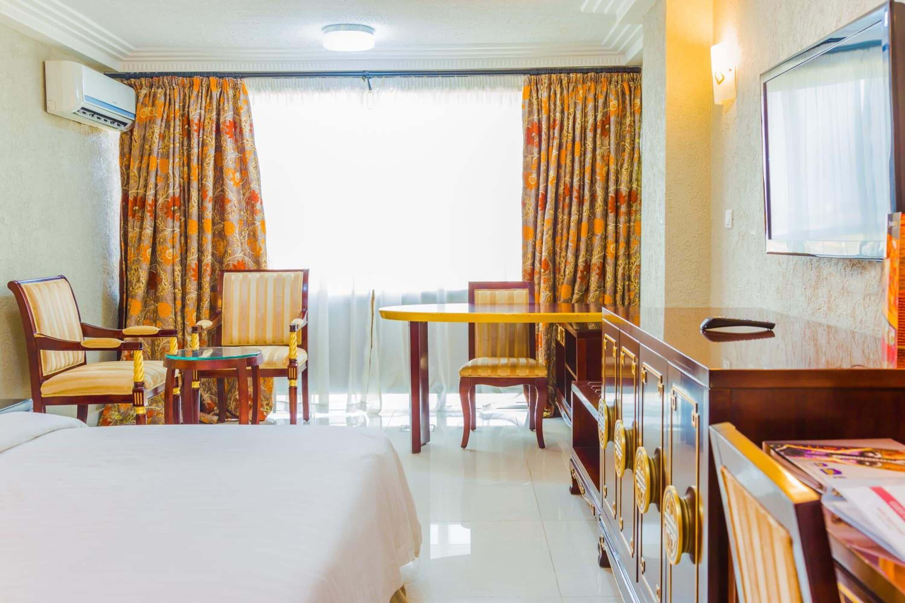 A hotel room with a bed, two chairs, a small table, a minibar, and a television.