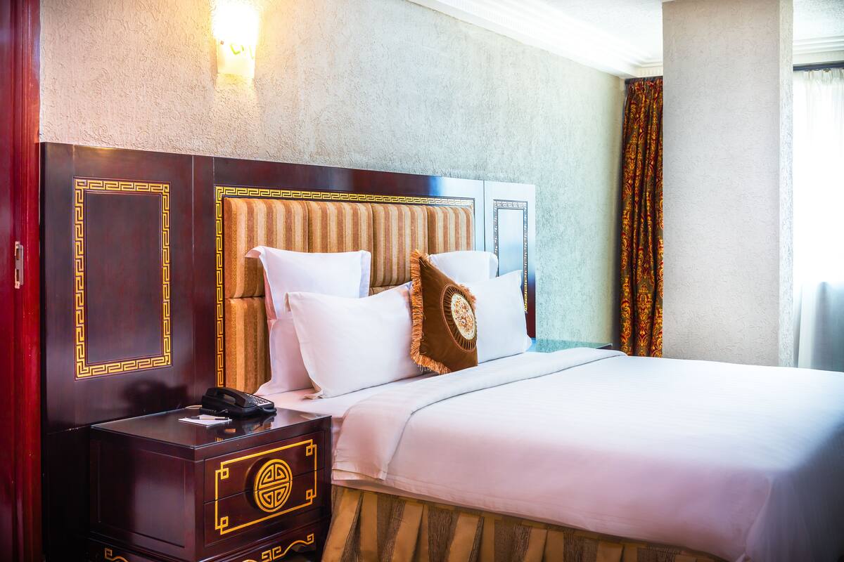 A hotel room with a large bed, a wooden nightstand with a telephone, and a wall-mounted light fixture.