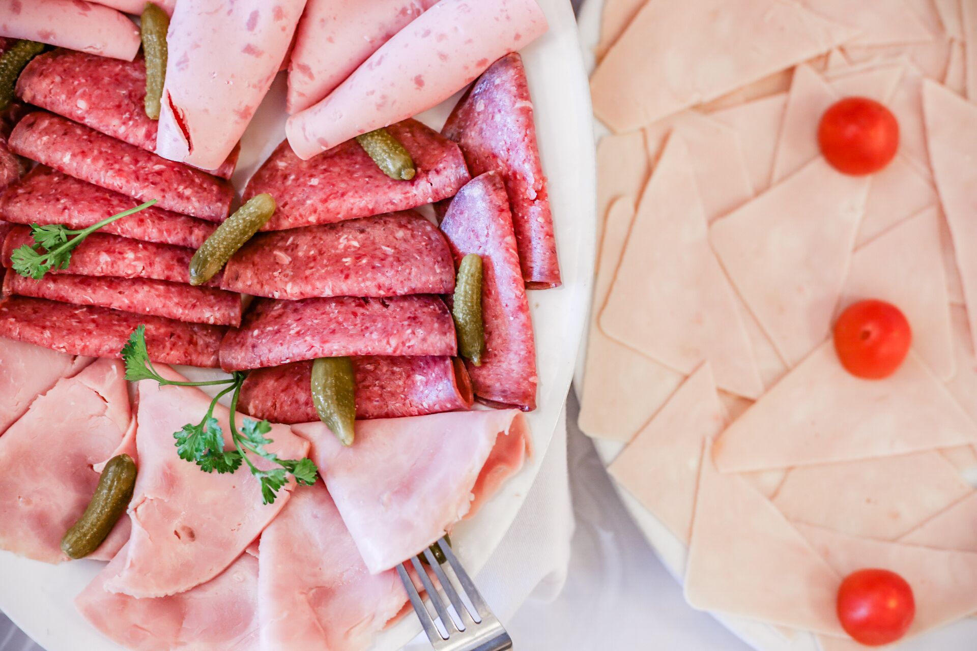 Un plato de embutidos con pepinillos y perejil, y otro plato de jamón con tomates cherry.