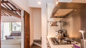 A kitchen with a gas stove, kettle, and shelves with kitchen items. There is a wooden door leading to another room with a sofa and striped cushions. - RSH Campo de Fiori Luxury House (Rome)
