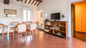 A dining area with a wooden table and chairs, a television on a wooden stand, and a bookshelf filled with books. - RSH Campo de Fiori Luxury House (Rome)