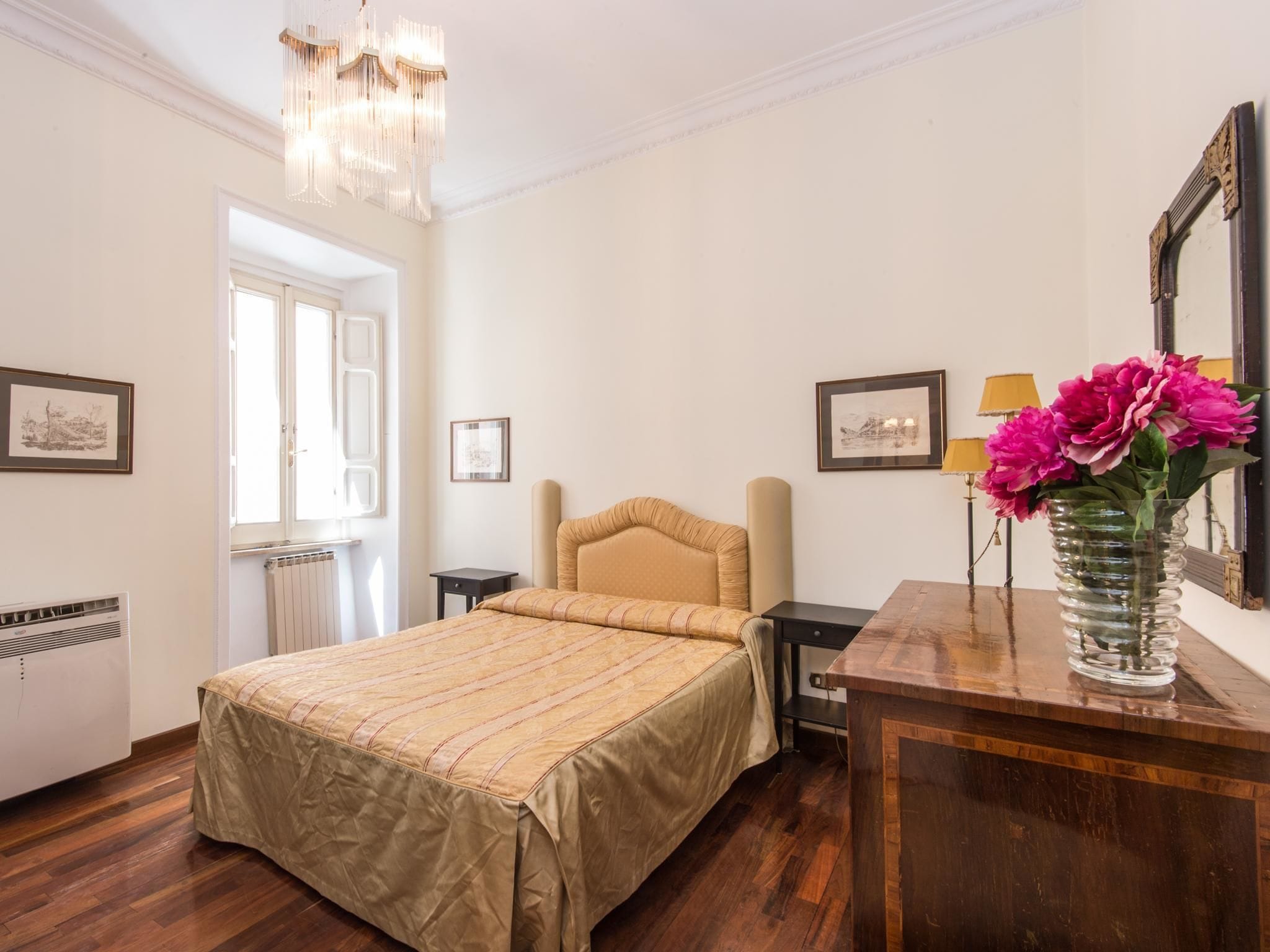A hotel room with a bed, a wooden dresser, a mirror, and a vase with flowers.