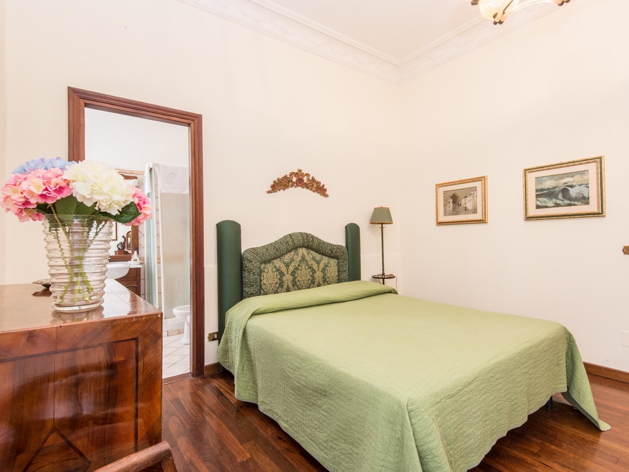 A bedroom with a bed, a dresser, a vase with flowers, a lamp, and two framed pictures on the wall.