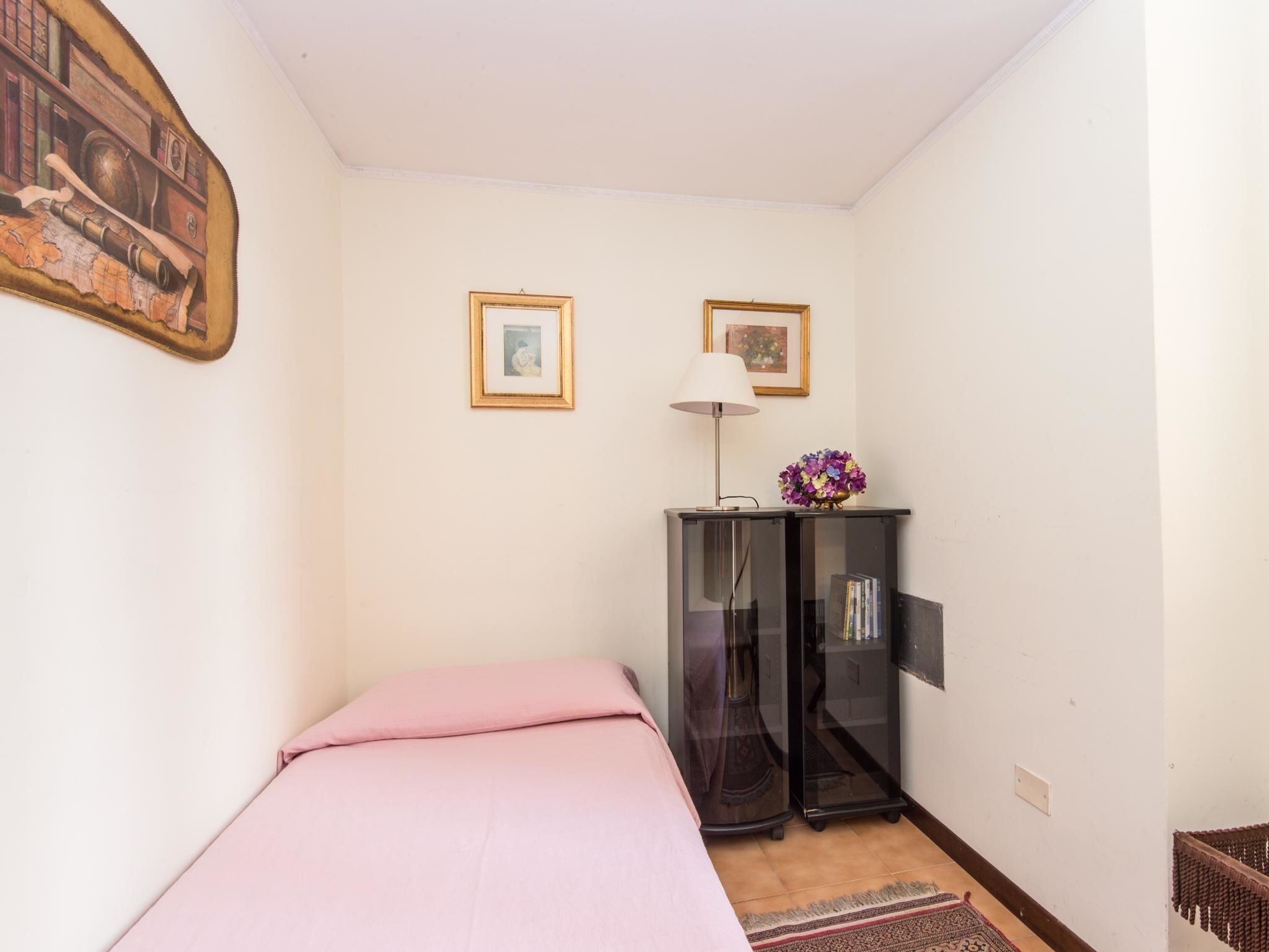 A small, neatly arranged bedroom with a single bed, a small cabinet, and two framed pictures on the wall.