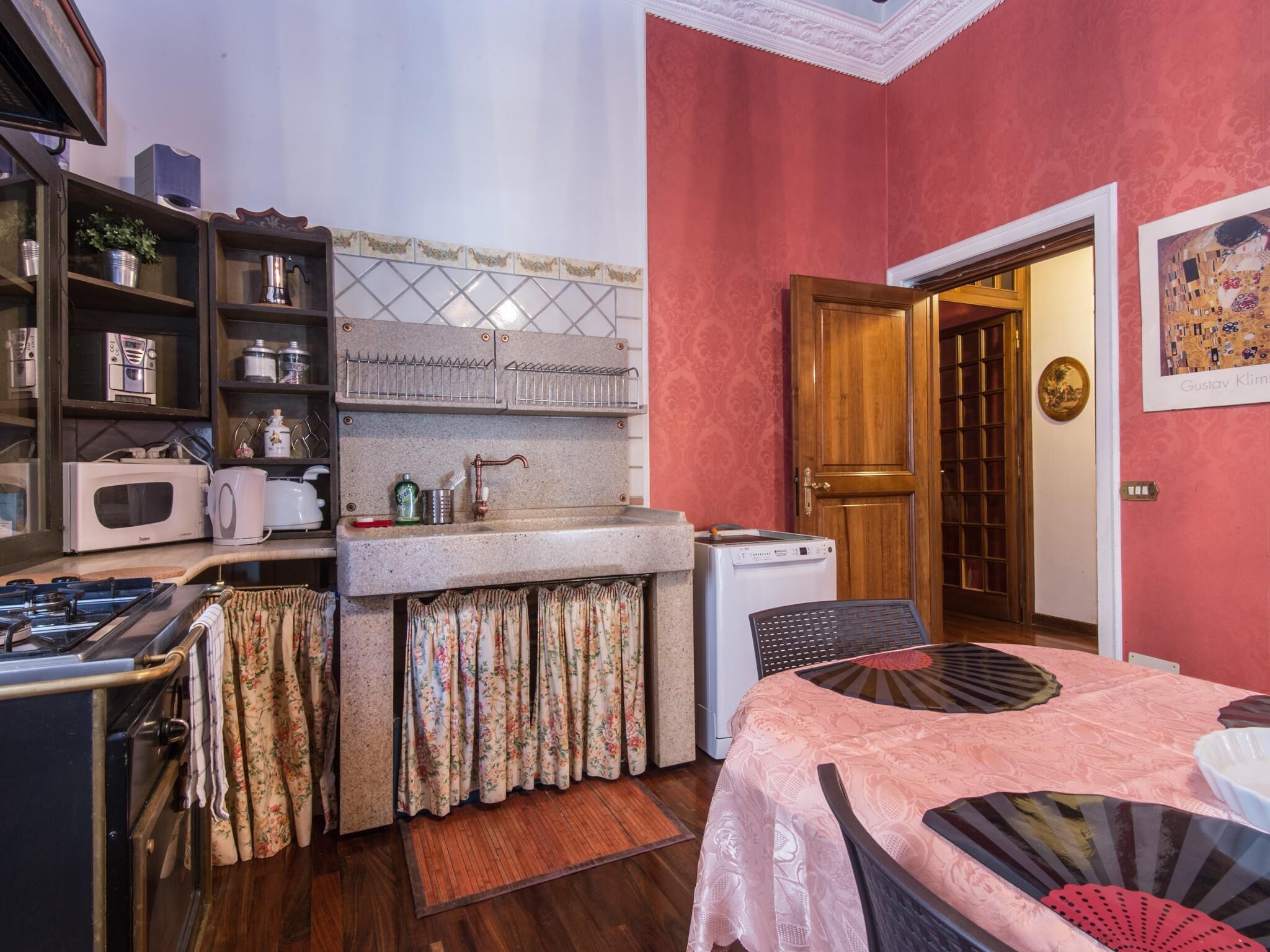 A compact kitchen with a microwave, toaster, and gas stove. There is a sink with a faucet, and a small refrigerator. The room has a wooden floor and a pink wall.