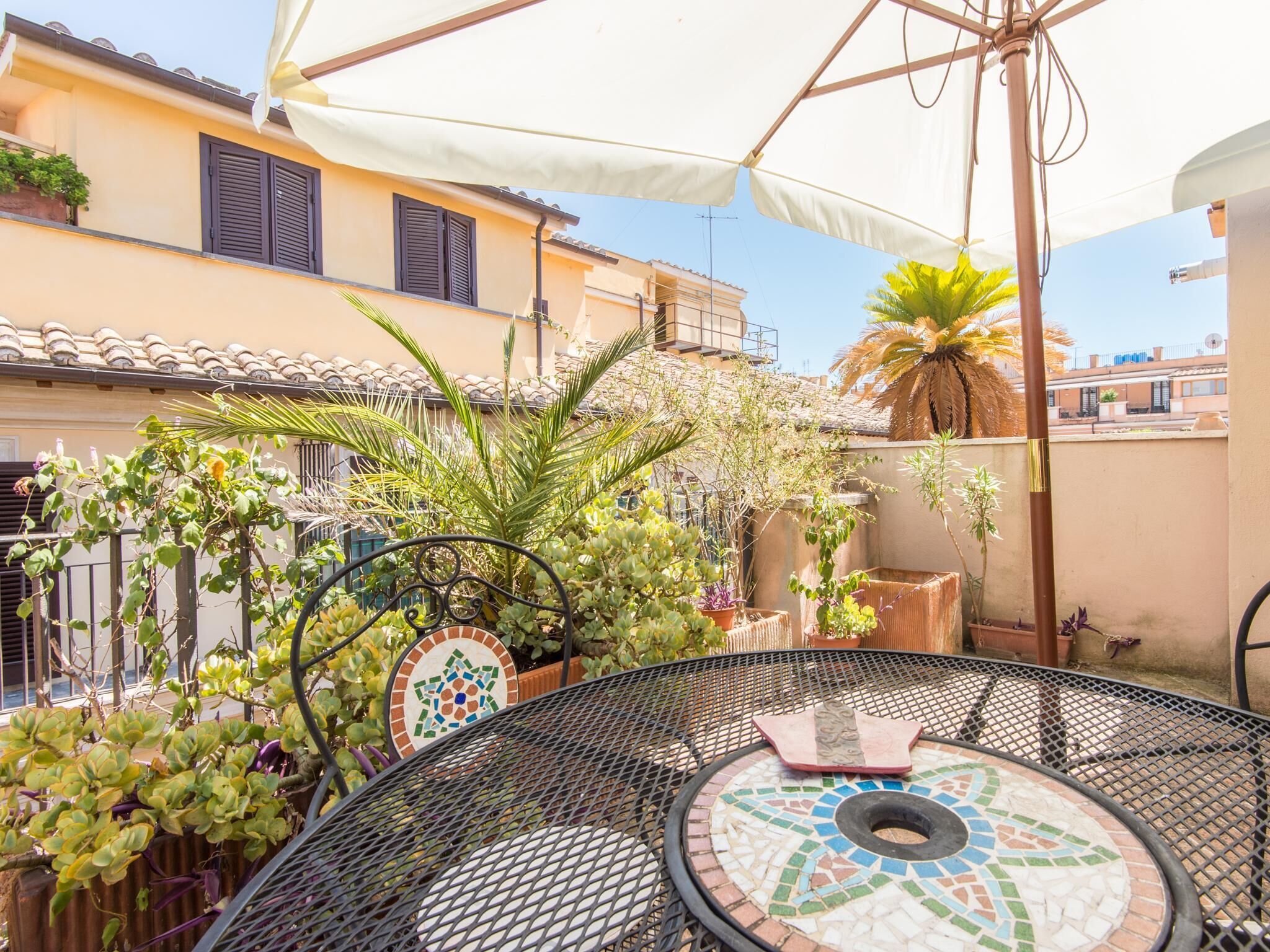A round metal table with a mosaic top, surrounded by potted plants and a white umbrella.