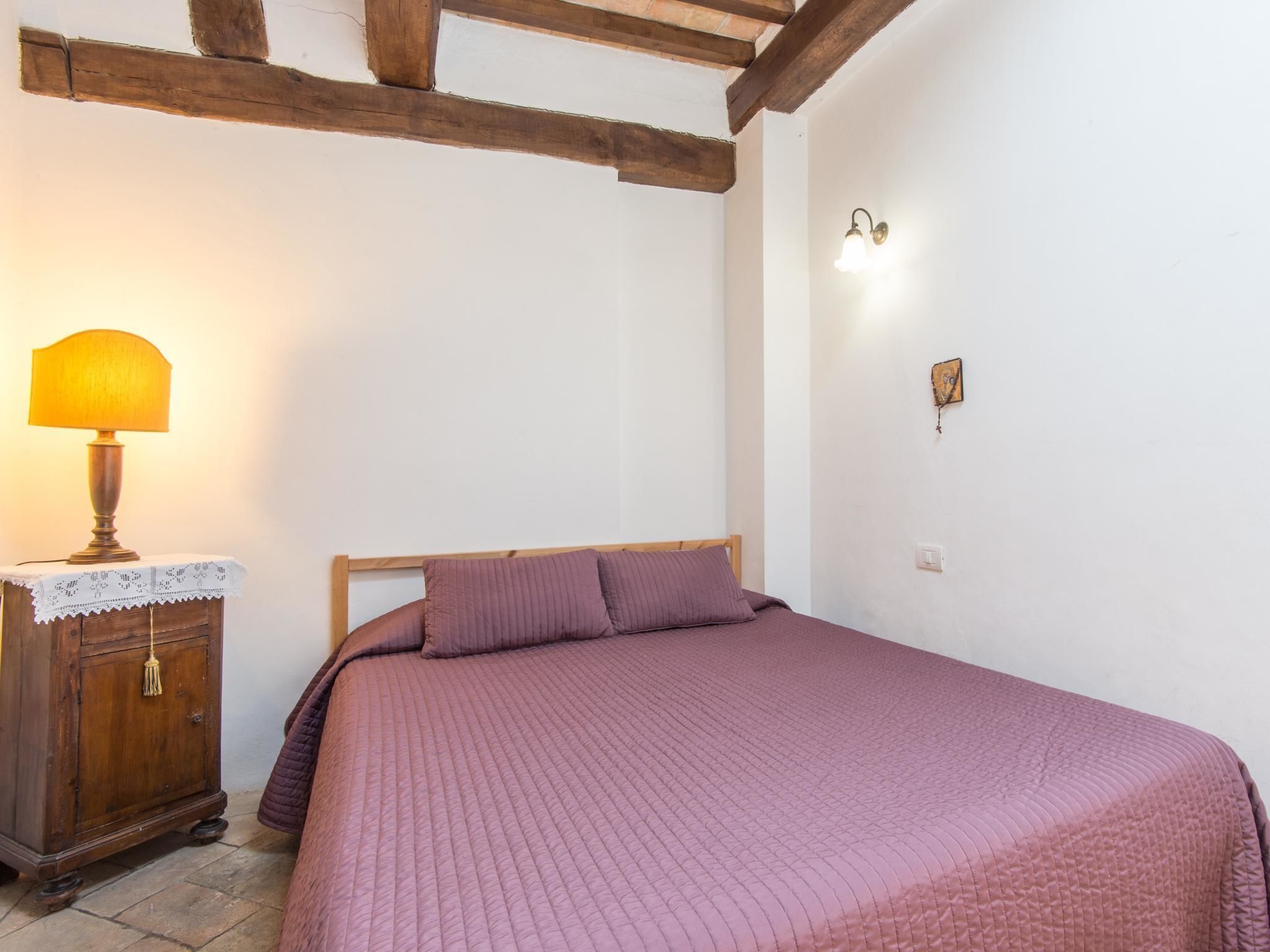 A bedroom with a wooden bed frame, a pink quilted bedspread, a wooden nightstand with a lamp, and a wall-mounted light fixture.
