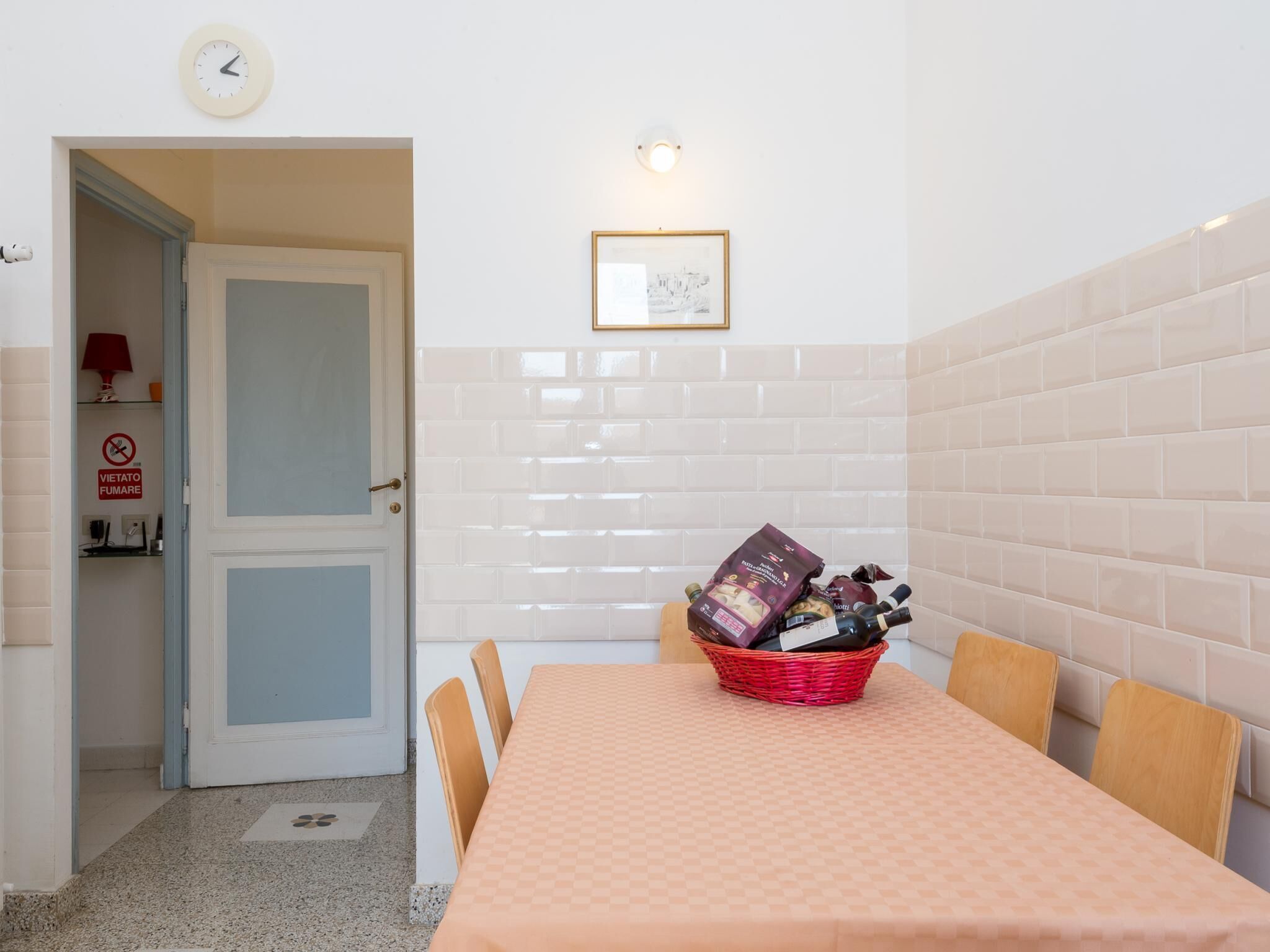 A dining area with a table set for four, a basket of wine bottles, a clock, and a wall-mounted light fixture.