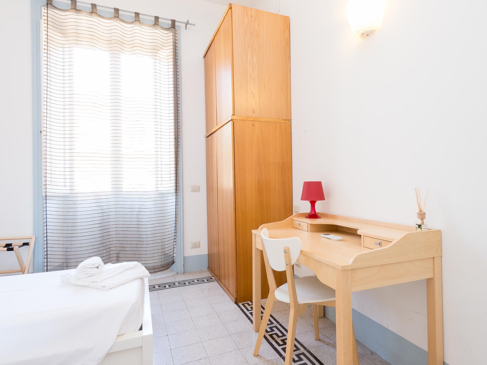 A small, well-lit room with a wooden desk, a white chair, a bed with a striped curtain, and a red lamp.