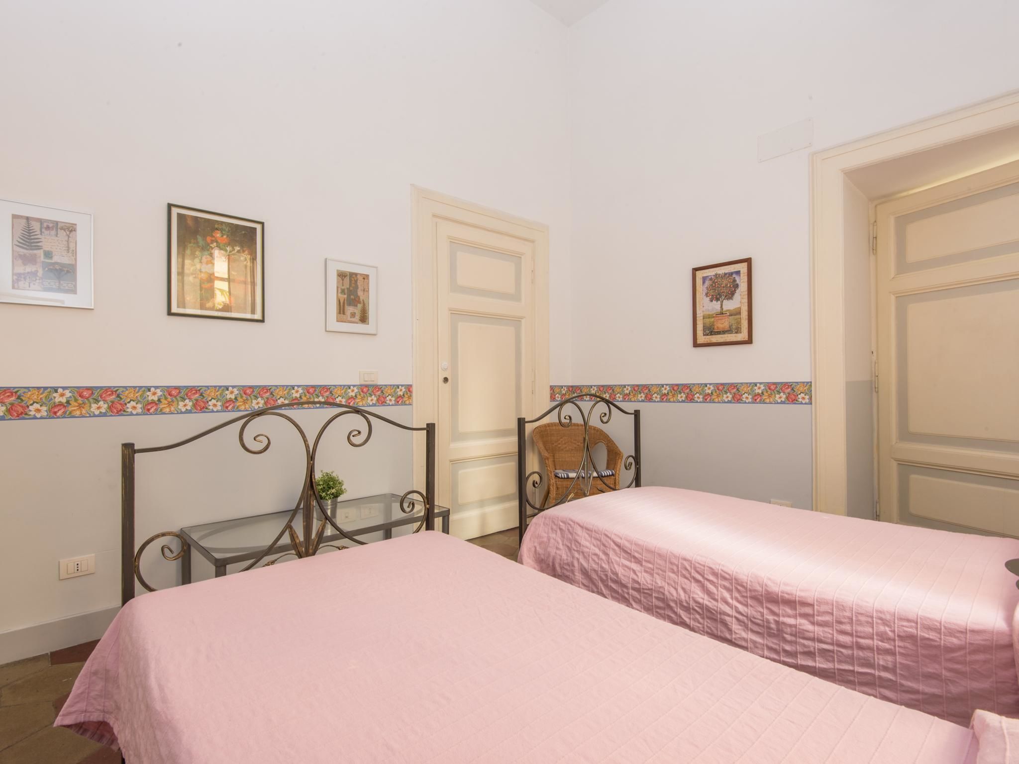 A bedroom with two beds, a small table, and a chair. There are doors and framed pictures on the walls.