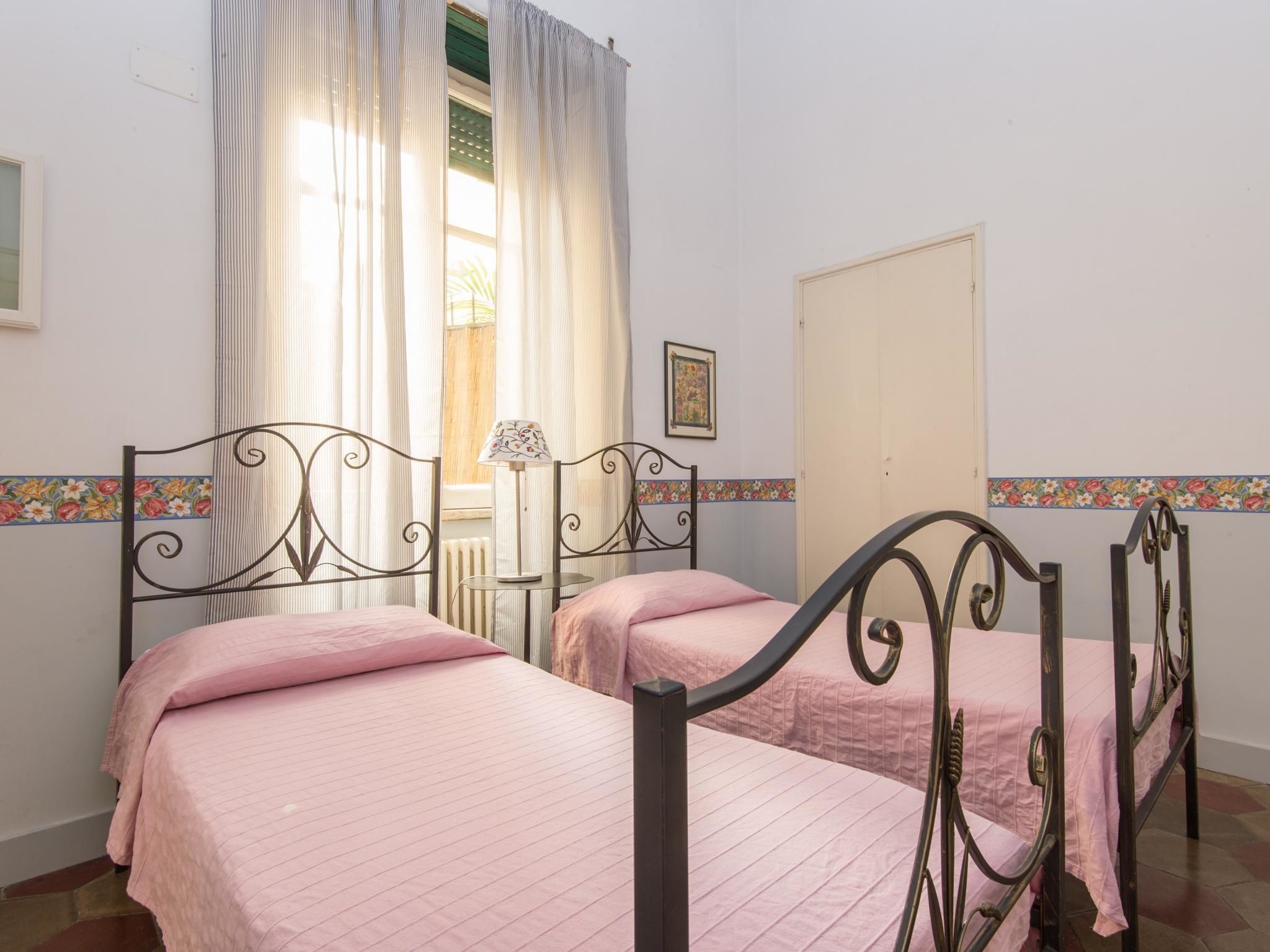 A room with two beds, each with a metal frame and pink bedding. There is a window with curtains, a radiator, and a door.