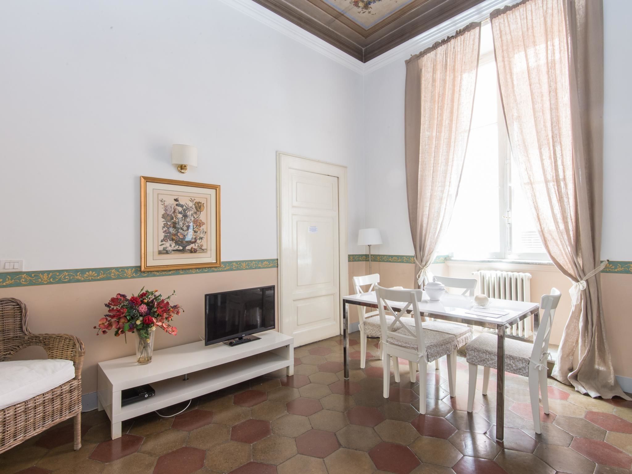 A room with a television on a white stand, a dining table with chairs, a wicker sofa, a vase with flowers, and a door with a light switch.