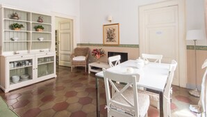 A dining area with a white table and chairs, a wicker chair, a cabinet with glass doors, and a painting on the wall. - RSH Ara Pacis Terrace Apartment (Rome)