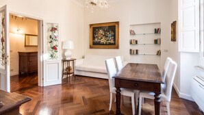 Ein Esszimmer mit Holztisch, weißen Stühlen, einer Chandelieber, einem Gemälde und einem Bücherregal.