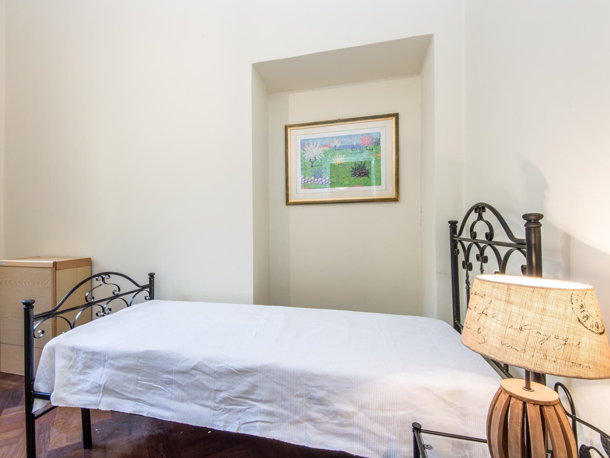 Un lit simple avec un drap blanc, une table de chevet en bois, une lampe et un tableau encadré au mur.