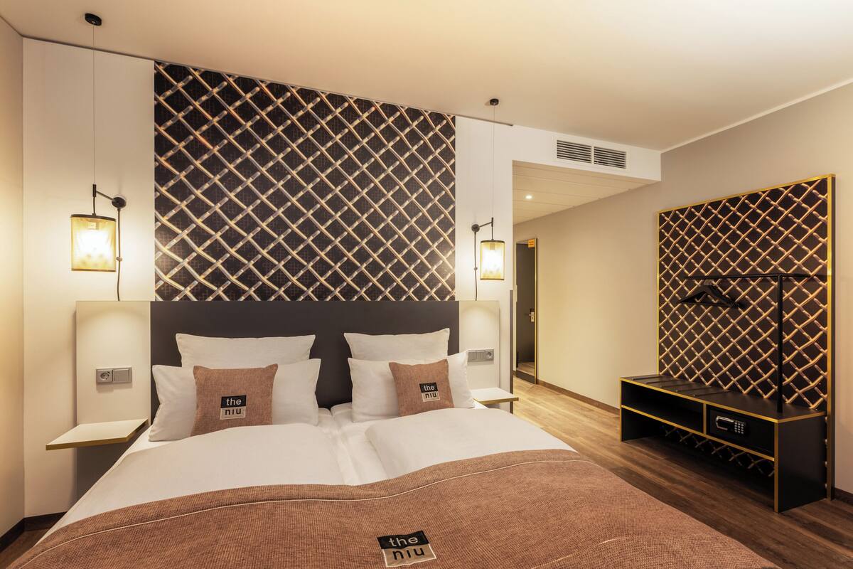 A modern hotel room with a large bed, two bedside tables, a built-in TV, and a minibar.