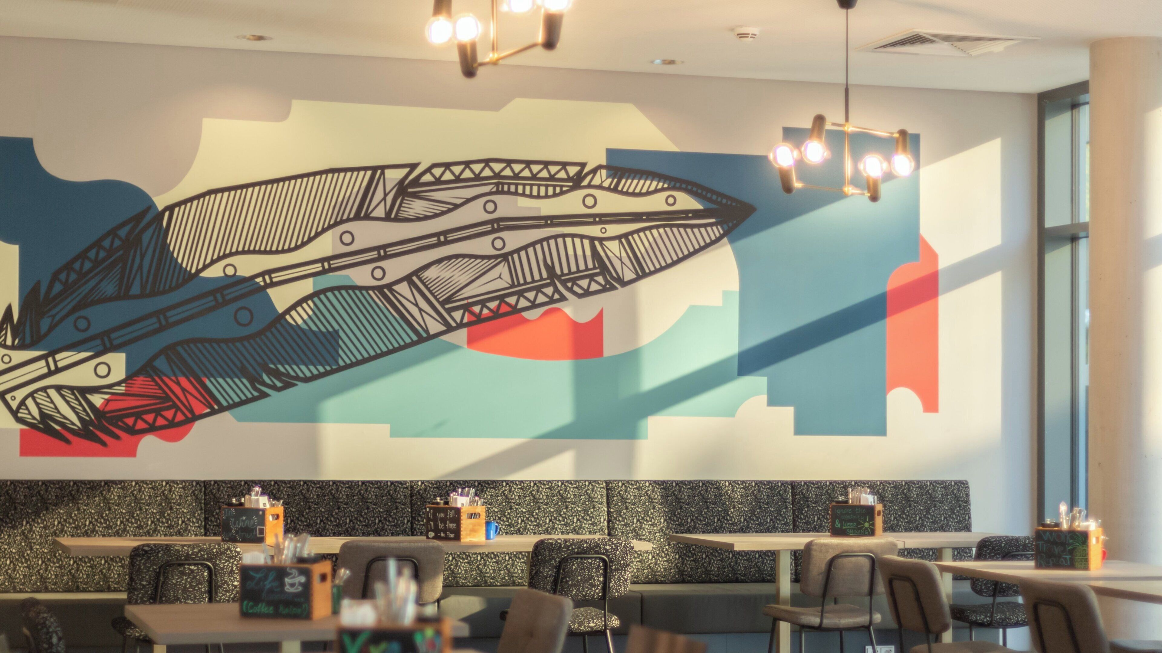 Interior de restaurante con un mural grande, asientos y una lámpara moderna.