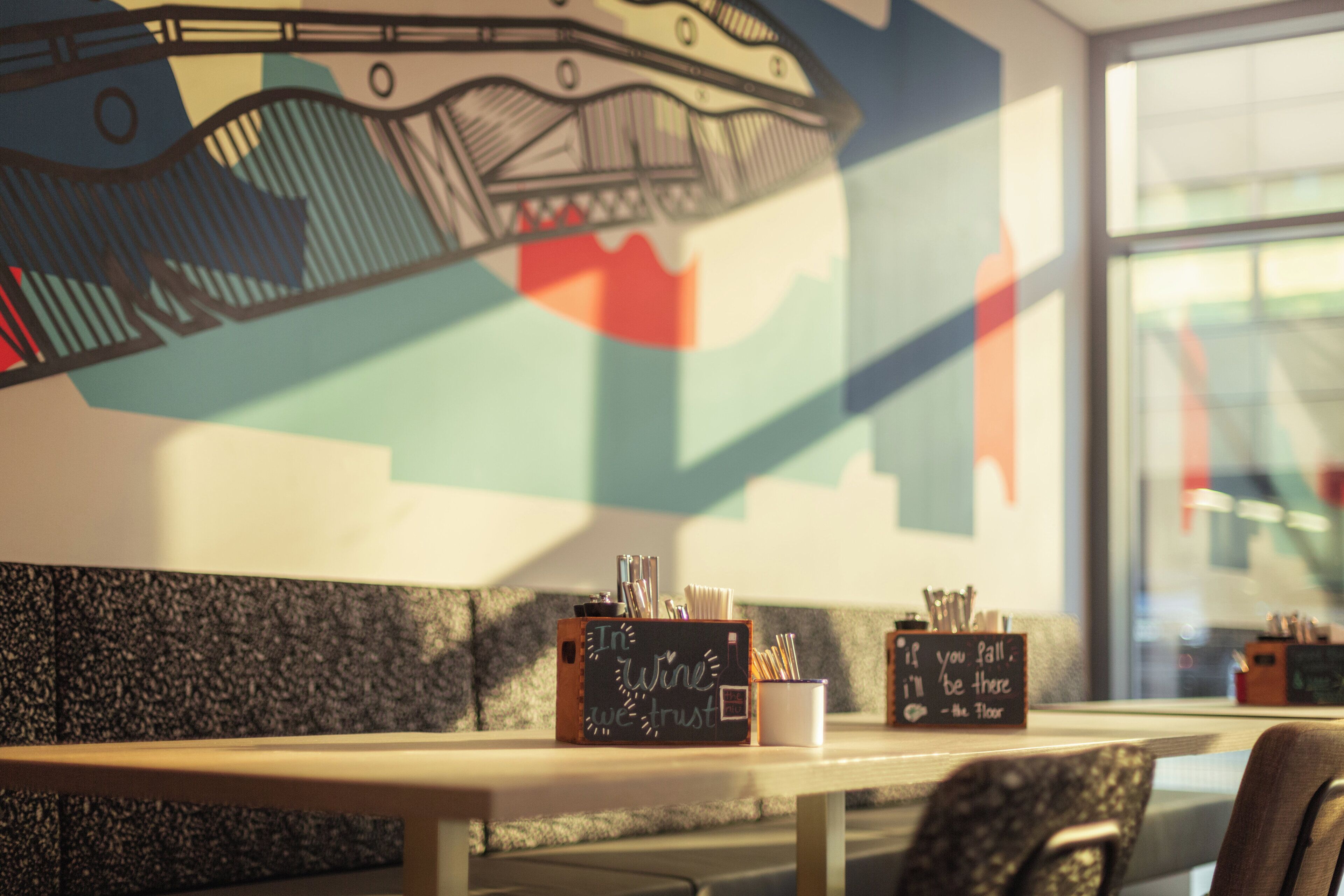 Una cafetería con un letrero de pizarra, una mesa, sillas y un mural en la pared.