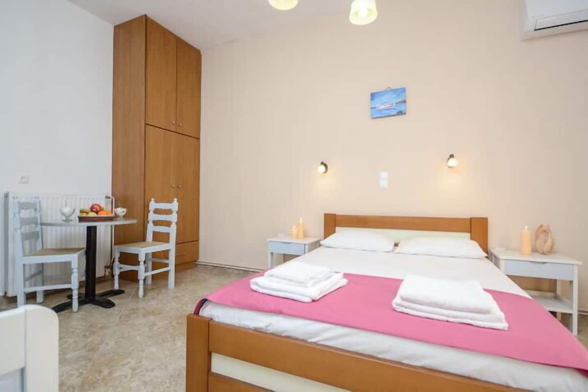 Quarto com cama, guarda-roupa de madeira, mesa pequena com cadeiras e luminária na parede.
