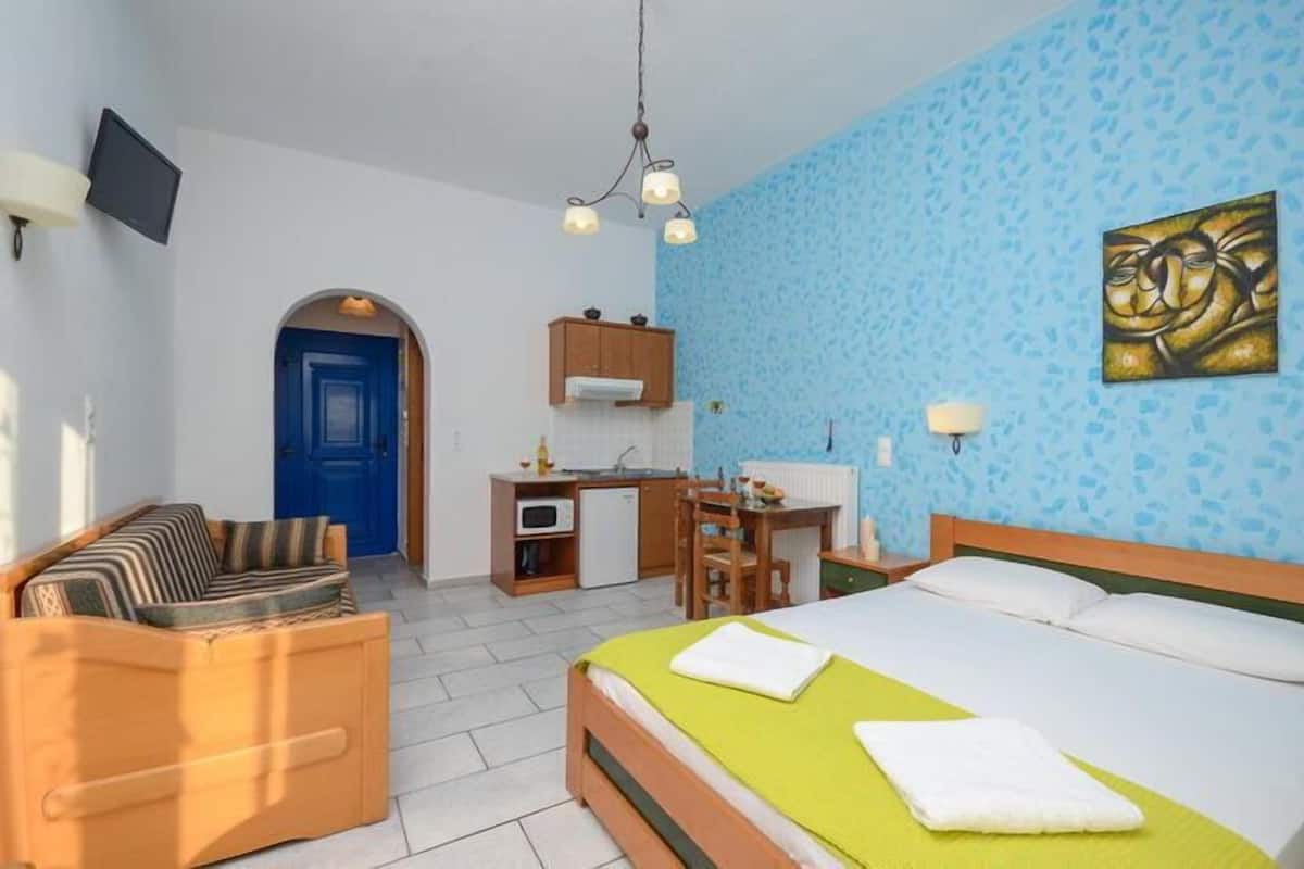 Quarto de hotel compacto com cama, sofá, escrivaninha com cadeira, cozinha compacta e uma porta azul.