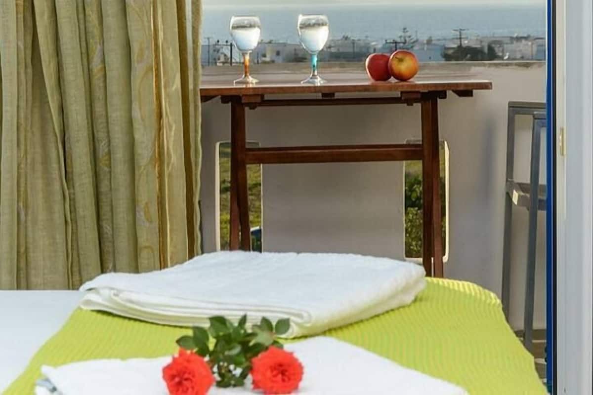 Cama bem arrumada com colcha verde, toalha branca e duas flores vermelhas. Ao fundo, vista para o mar e uma mesa com dois copos e uma maçã.