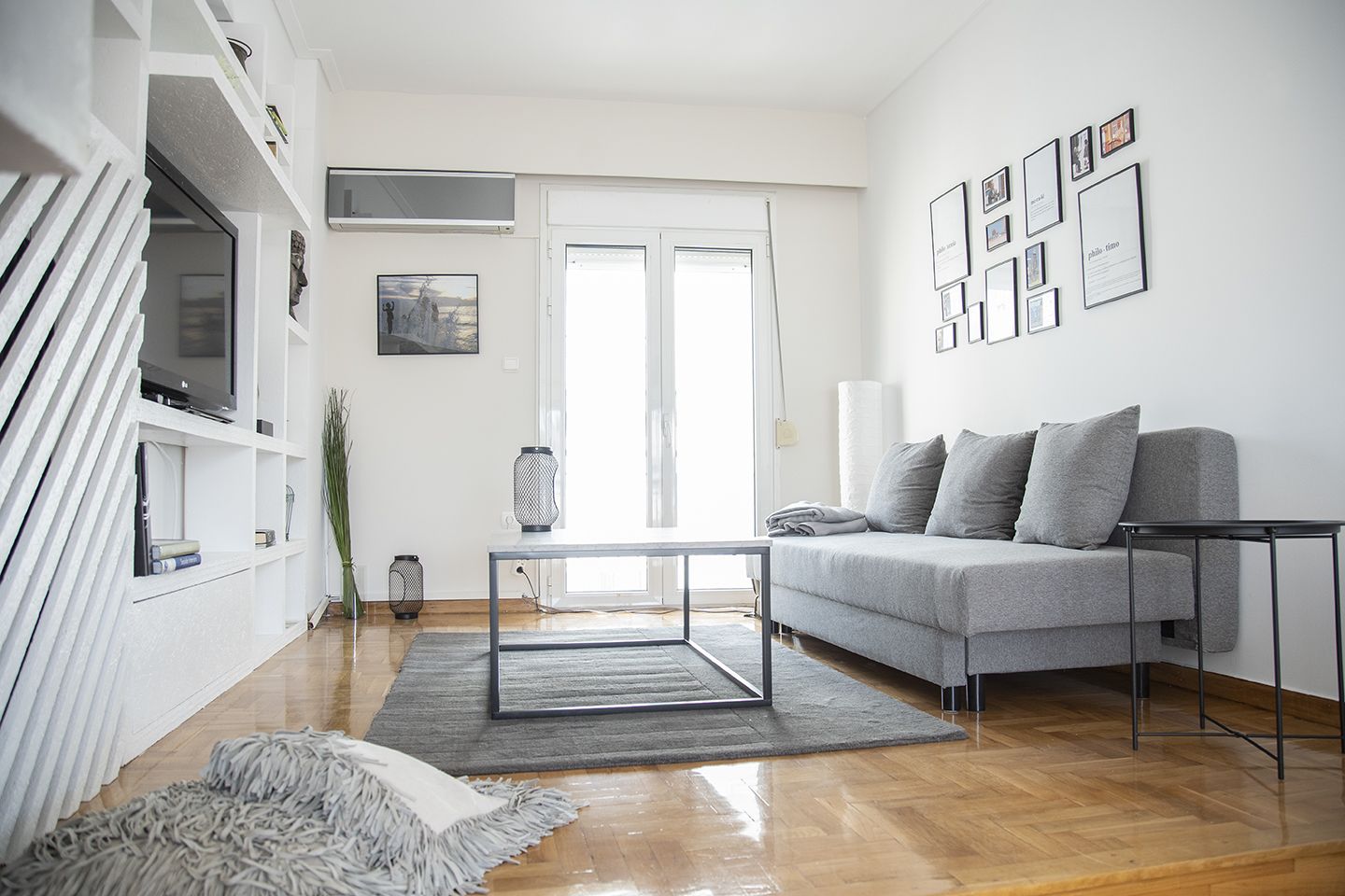 A modern living room with a grey sofa, a coffee table, and a TV on a white wall. There are framed pictures and a plant on the wall.